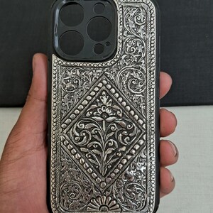 Puede incluir: Una funda de tel&eacute;fono plateada con intrincados dise&ntilde;os florales. La funda est&aacute; hecha de metal y tiene un dise&ntilde;o en relieve. La funda est&aacute; dise&ntilde;ada para un iPhone 12 Pro Max.
