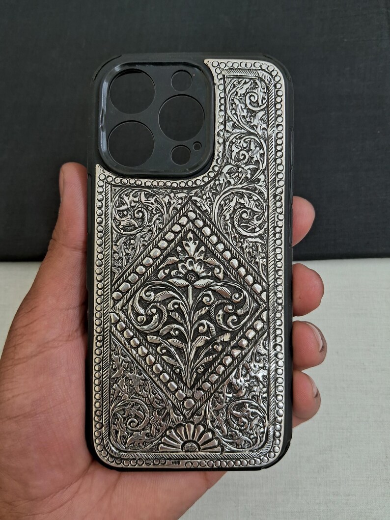 Puede incluir: Una funda de tel&eacute;fono plateada con intrincados dise&ntilde;os florales. La funda est&aacute; hecha de metal y tiene un dise&ntilde;o en relieve. La funda est&aacute; dise&ntilde;ada para un iPhone 12 Pro Max.