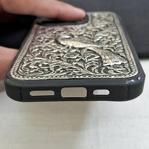 以下が含まれることがあります： 銀色の複雑な花柄が施された黒い携帯電話ケース。ケースはカメラレンズの周りに盛り上がった縁があり、充電ポート用の切り抜きがあります。