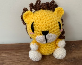 Puede incluir: Un león amarillo de crochet con pelo marrón, patas blancas y ojos negros.
