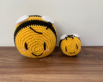 Puede incluir: Dos juguetes con forma de abeja hechos a ganchillo. La abeja más grande es amarilla con rayas negras y alas blancas. La abeja más pequeña tiene el mismo diseño. Ambos tienen caras sonrientes con mejillas rosadas. Los juguetes están sobre una superficie de madera.