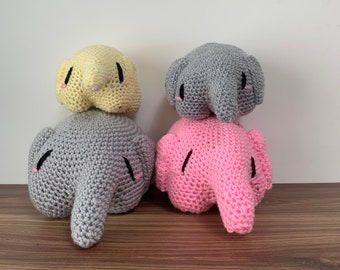 Puede incluir: Cuatro elefantes de peluche tejidos a crochet, uno amarillo, uno rosa y dos grises, apilados uno encima del otro. Los elefantes tienen ojos negros y mejillas rosadas.