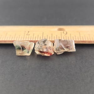 May include: Three small, clear, faceted sunstone crystals with a slight green, red, and orange hue. The crystals are arranged in a row on a dark surface.