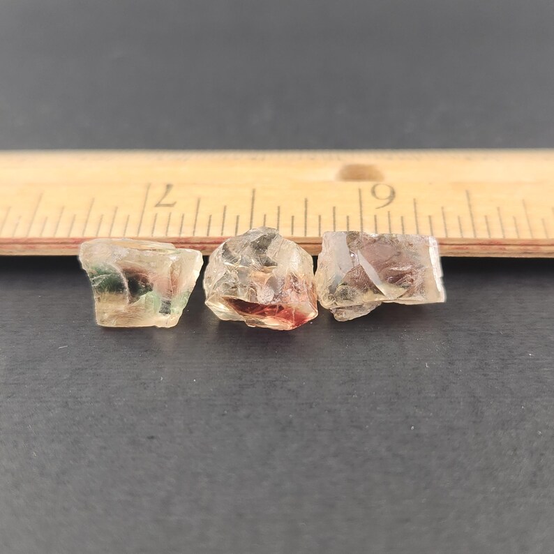 May include: Three small, clear, faceted sunstone crystals with a slight green, red, and orange hue. The crystals are arranged in a row on a dark surface.