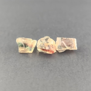 May include: Three small, translucent, faceted gemstones with a variety of colors, including green, red, and orange. The gemstones are arranged in a row on a gray surface.