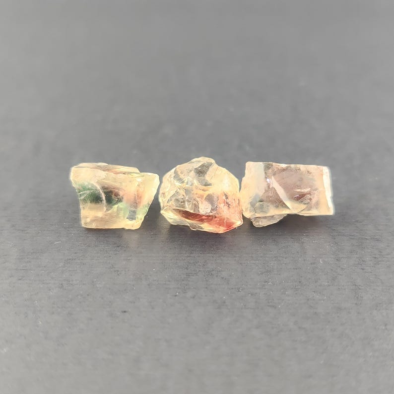 May include: Three small, translucent, faceted gemstones with a variety of colors, including green, red, and orange. The gemstones are arranged in a row on a gray surface.