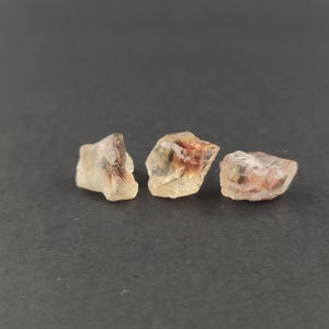 May include: Three rough, translucent sunstone crystals with a golden-yellow hue and reddish-brown inclusions. The crystals are arranged in a row on a dark gray surface.