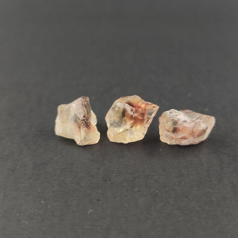 May include: Three rough, translucent sunstone crystals with a golden-yellow hue and reddish-brown inclusions. The crystals are arranged in a row on a dark gray surface.