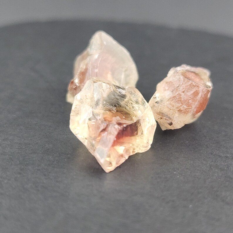 May include: Three sunstone crystals, two are clear with a reddish-orange hue and one is a lighter yellow with a reddish-orange hue.