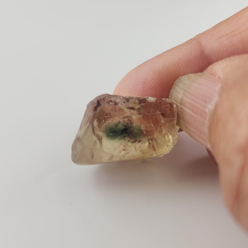 Op de afbeelding: Een doorschijnende geelgroene sunstone edelsteen met een groene insluiting. De steen is onregelmatig gevormt en heeft een ruwe, natuurlijke afwerking.