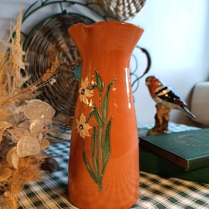 Peut inclure: Un vase en céramique orange avec un bord évasé, orné d'un motif peint à la main de jonquilles blanches et de feuilles vertes. Le vase est posé sur une nappe à carreaux verts et blancs, avec des objets décoratifs en arrière-plan.