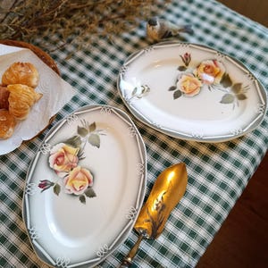 Peut inclure: Deux plats de service ovales avec un motif de roses jaunes et roses. Les plats ont un fond blanc avec une bordure grise et des accents blancs décoratifs. Une pelle à gâteau dorée est également visible.