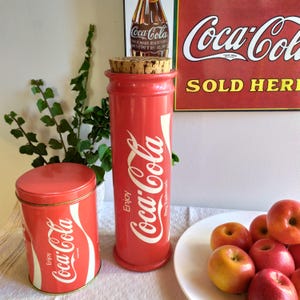 Peut inclure: Deux contenants Coca-Cola rouges, l'un un cylindre haut avec un bouchon en liège et l'autre une boîte de conserve plus courte. Les deux présentent le logo Coca-Cola et la phrase "Enjoy".