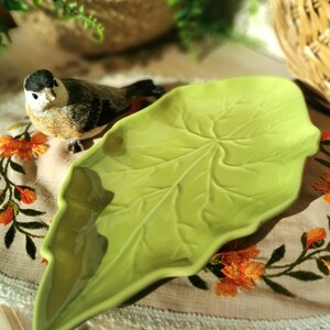 Peut inclure: Un plat de service en céramique vert clair en forme de feuille. Le plat présente des nervures détaillées et une finition brillante. Une figurine décorative d'oiseau est positionnée près du plat. Le fond comprend un panier tressé et des détails floraux brodés.