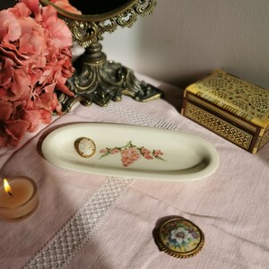 Peut inclure: Plat ovale en céramique blanche avec un motif floral et une petite broche camée sur le dessus. Le plat est sur une nappe blanche avec une petite boîte et une bougie en arrière-plan.