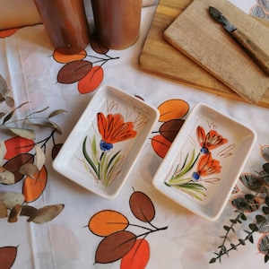 Peut inclure: Deux plats rectangulaires en céramique blanche avec des motifs floraux orange et bleus. Les plats sont sur une nappe blanche avec un motif de feuilles brunes et orange.
