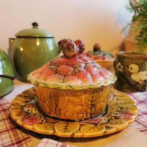 Peut inclure: Un beurrier en céramique avec un couvercle en forme de panier de fraises. Le plat est décoré de feuilles vertes et de fraises rouges. Le couvercle a deux petits oiseaux perchés sur le dessus.