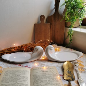 Peut inclure: Deux plats de service en céramique blanche en forme de cygne sur une table avec un livre et un ustensile de service doré. Les plats sont sur une nappe blanche à motifs brun et jaune.