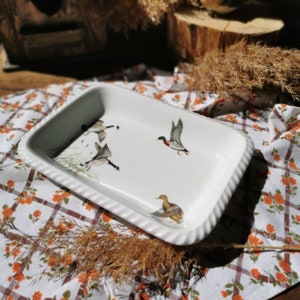 Peut inclure: Plat à four en céramique blanche avec un bord surélevé et un motif de canard et d'oies. Le plat est sur une nappe à carreaux blancs et bruns.