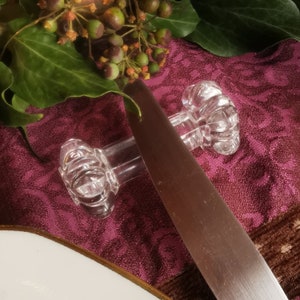 Peut inclure: Ronds de serviette en verre transparent avec un design côtelé, tenant un couteau en acier inoxydable. Le couteau repose sur une nappe bordeaux à motifs floraux. Feuilles vertes et baies en arrière-plan.