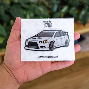 Peut inclure: Patch brodé blanc représentant une voiture argentée avec des détails noirs et des phares jaunes. Le patch comporte le texte "Run the Streets" au-dessus de la voiture et "@runthestreets.shop" en dessous.