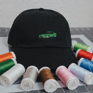 May include: A black baseball cap with a green embroidered car on the front. The cap is surrounded by spools of colorful thread.