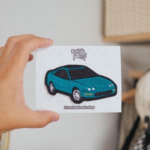 May include: Embroidered patch featuring a teal coupe car with black roof and white wheels. The patch has the text "RUN THE STREETS" and "@runthestreets.shop". The patch is held in a hand.