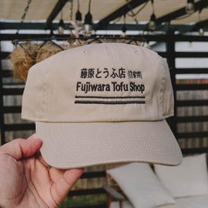 Op de afbeelding: Een beige honkbalpet met zwarte geborduurde tekst die Fujiwara Tofu Shop leest, samen met Japanse karakters en twee horizontale lijnen. De pet heeft een gestructureerde kroon, een gebogen klep en gestikte oogjes op de bovenste panelen.