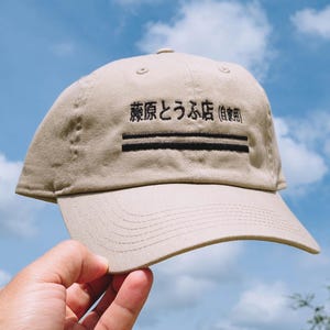 May include: A beige baseball cap featuring black embroidered Japanese text that reads 藤原とうふ店 (自家用) and two horizontal black lines centred on the front panel. The cap has a structured crown, a curved brim with stitching, and a button on top.