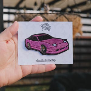 May include: Embroidered patch depicting a pink sports car with black detailing and yellow wheels. The patch is on a white card with the text "Run the Streets" and "@runthestreets.shop".