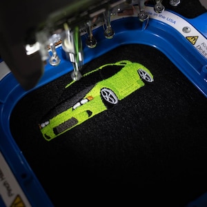 May include: An embroidered design of a lime green sports car with black accents and white wheels. The car is being stitched onto black fabric by an embroidery machine. The machine is blue and has the text "Made in the USA".