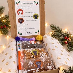 May include: A brown cardboard Christmas Eve box containing three clear plastic bags of cat treats, a purple fuzzy toy, and a gold glitter ball. The box lid features a printed list of contents including Webbox Lick-e-Lix and crunchy treats.