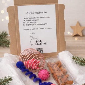 May include: A brown cardboard box containing a cat toy set. The label reads Purrfect Playtime Set, listing a coil spring toy with a rattle mouse, a sparkle pompom, a cool pompom, and 20g of chicken cushions for adult cats and kittens 2+ months.