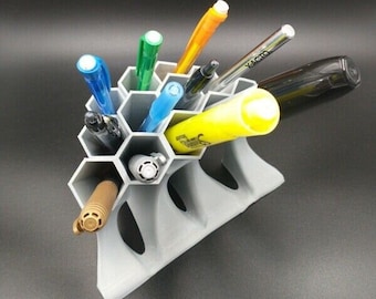 Op de afbeelding: Een grijze, zeshoekige pennenhouder van plastic met een honingraatontwerp. De houder is gevuld met verschillende pennen, waaronder een zwarte pen, een blauwe pen, een groene pen, een gele markeerstift, een oranje pen en een bruine pen.
