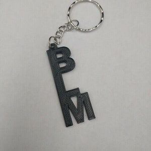 Peut inclure: Porte-clés noir avec les lettres "BLM" imprimées dessus. Le porte-clés est attaché à un anneau en métal argenté.