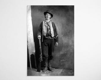 May include: A black and white historical photograph of a person wearing a wide-brimmed hat, a patterned neckerchief, a buttoned vest over a dark shirt, trousers, and tall boots. The person stands holding a long rifle in a vertical position.