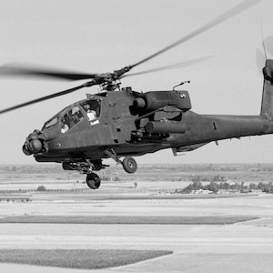 May include: A black and gray military helicopter in flight over a field. The helicopter has a large rotor blade and a tail rotor.