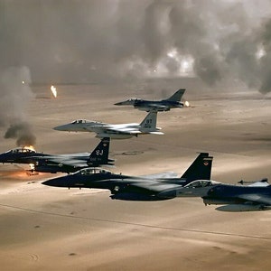 May include: A group of four fighter jets fly over a desert landscape with smoke and flames in the background. The jets are all in formation and appear to be in flight. The sky is cloudy and the sun is shining.