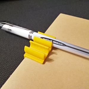 K&ouml;nnte beinhalten: Ein gelber Plastik-Stiftst&auml;nder mit einem wei&szlig;en Stift mit schwarzen Akzenten und dem Text "Uniball Vision Elite" darauf.
