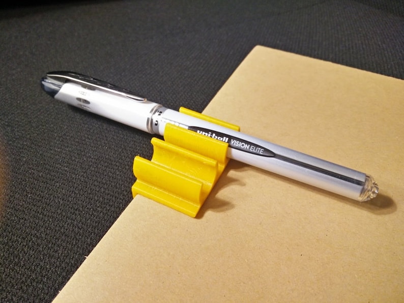 K&ouml;nnte beinhalten: Ein gelber Plastik-Stiftst&auml;nder mit einem wei&szlig;en Stift mit schwarzen Akzenten und dem Text "Uniball Vision Elite" darauf.