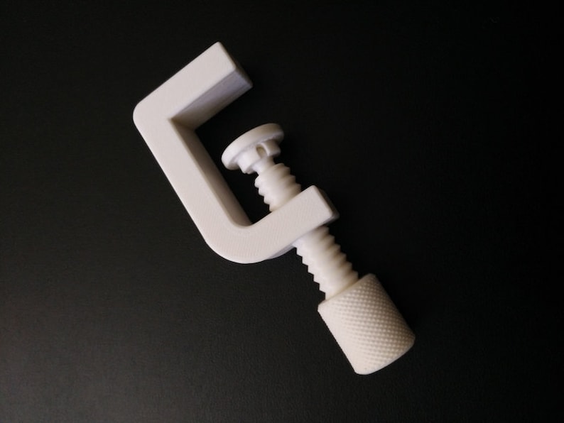 May include: A white plastic clamp with a threaded screw. The clamp is open and the screw is partially turned.