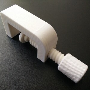May include: A white plastic clamp with a threaded screw and a knob. The clamp is designed to hold objects together.