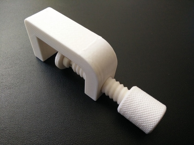 May include: A white plastic clamp with a threaded screw and a knob. The clamp is designed to hold objects together.