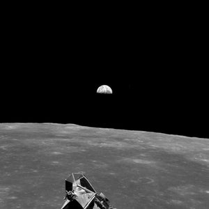 May include: Black and white photograph of the lunar surface with the Earth visible in the distance. A lunar module is in the foreground, with the Earth rising over the horizon. The image is taken from a low angle.