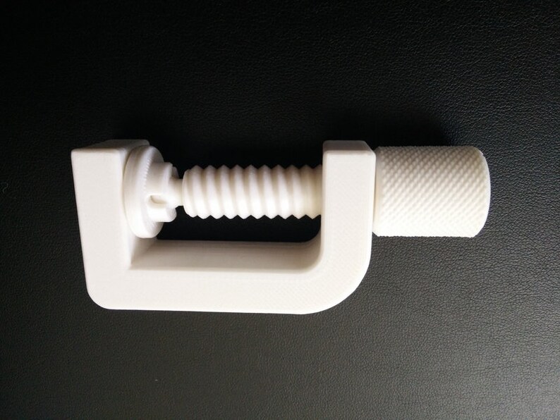 May include: A white plastic clamp with a threaded screw and a knob for tightening. The clamp is designed to hold objects together.
