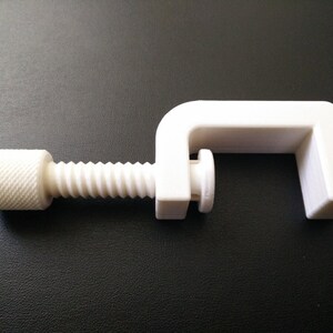 May include: A white plastic clamp with a threaded screw. The clamp is designed to hold objects together.