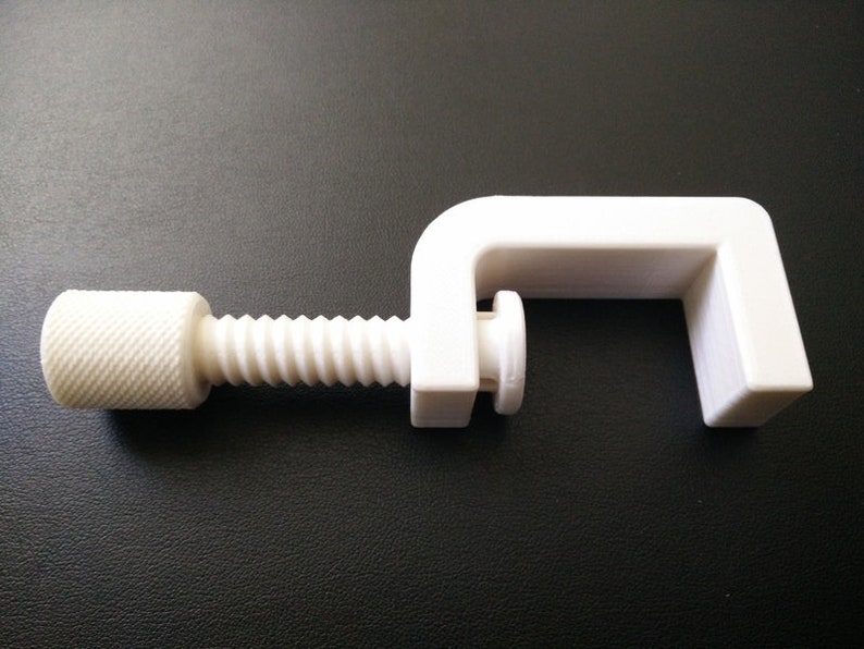 May include: A white plastic clamp with a threaded screw. The clamp is designed to hold objects together.