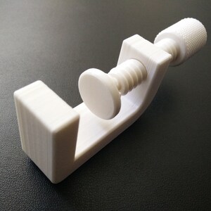 May include: A white plastic clamp with a threaded screw and a knob. The clamp is open and ready to hold an object.