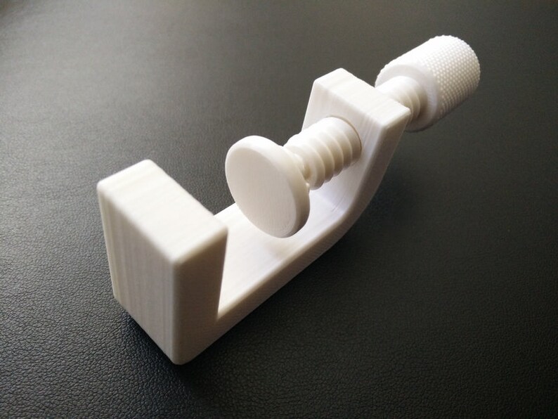 May include: A white plastic clamp with a threaded screw and a knob. The clamp is open and ready to hold an object.