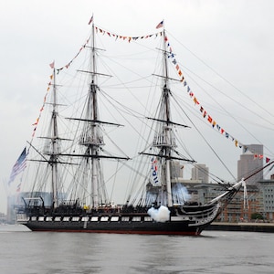 May include: A black and white sailing ship with a white smoke plume coming from the cannon on the deck. The ship is decorated with flags and is sailing in a harbor with a city skyline in the background.